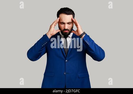 Homme barbu qui frognait et attrape la tête, souffrant de maux de tête intenses, ayant des symptômes insupportables de migraine, de fièvre et de grippe, portant un costume de style officiel. Prise de vue en studio isolée sur fond gris Banque D'Images