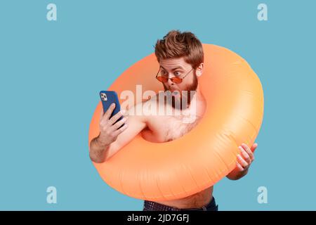 Portrait d'un homme à barbe surpris avec lunettes de soleil debout avec anneau en caoutchouc orange et tenant le téléphone cellulaire, regardant l'affichage avec la bouche ouverte. Studio d'intérieur isolé sur fond bleu. Banque D'Images