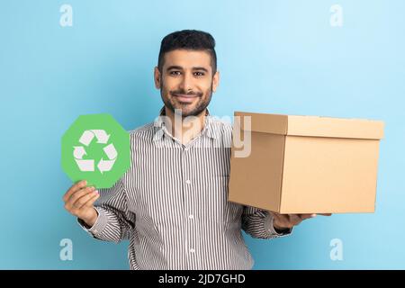 Homme responsable et confiant tenant l'affiche de recyclage verte dans la main et l'emballage en carton, regardant sourire à l'appareil photo, portant une chemise rayée. Studio d'intérieur isolé sur fond bleu. Banque D'Images