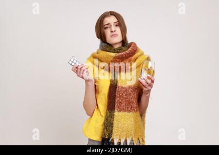 Portrait d'une adolescente malade aux cheveux bouclés enveloppée dans un foulard qui frotte le visage, tenant un verre d'eau et de pilules, le traitement, regardant l'appareil photo. Prise de vue en studio isolée sur fond gris. Banque D'Images