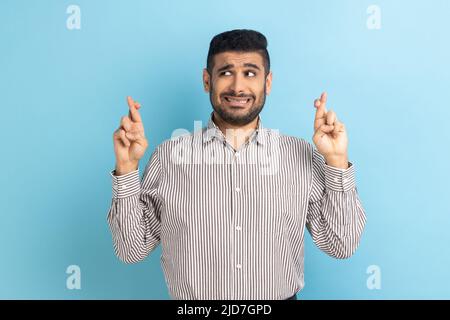 Portrait d'un jeune homme d'affaires plein d'espoir, debout avec un doigt croisé, des dents accroclées et l'espoir de gagner, portant une chemise rayée. Studio d'intérieur isolé sur fond bleu. Banque D'Images