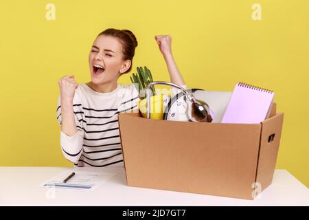 Portrait d'une heureuse femme excitée assise sur une table avec une boîte avec ses affaires personnelles, se réjouissant d'obtenir un nouvel emploi, exprimant des émotions positives. Studio d'intérieur tourné isolé sur fond jaune. Banque D'Images