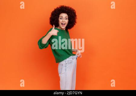 Portrait d'une femme positive excitée avec une coiffure afro portant un pull vert décontracté avec une taille mince dans un grand pantalon et des pouces vers le haut. Studio d'intérieur isolé sur fond orange. Banque D'Images