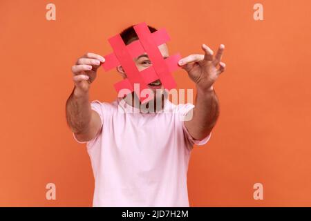 Portrait d'un homme barbu couvrant le visage avec le symbole hashtag des médias sociaux, recommandant de suivre le contenu tendance, blog populaire, portant un T-shirt rose. Studio d'intérieur isolé sur fond orange. Banque D'Images