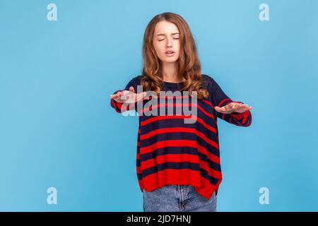 Portrait d'une femme solitaire aveugle et handicapée portant un chandail rayé, debout avec les yeux fermés et les mains tendues cherchant le chemin, marchant avec des doutes. Studio d'intérieur isolé sur fond bleu. Banque D'Images