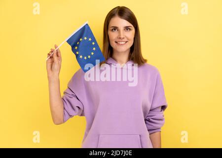 Femme patriotique aux cheveux foncés tenant le drapeau de l'ue à la main regardant l'appareil photo avec un sourire goûtant, patriotisme, célébration du jour du drapeau, portant le sweat à capuche violet. Studio d'intérieur isolé sur fond jaune. Banque D'Images