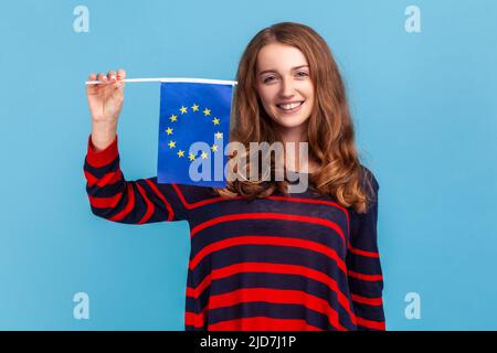 Femme optimiste portant un pull rayé de style décontracté, montrant le drapeau de l'Union européenne, regardant la caméra avec le sourire, l'association et la communauté de l'UE. Studio d'intérieur isolé sur fond bleu. Banque D'Images