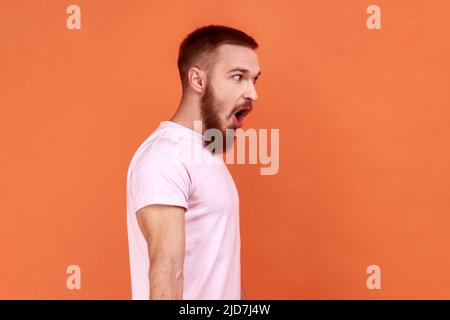 Vue latérale de l'homme barbu stupéfié debout avec la bouche ouverte dans la surprise, a choqué l'expression, entend des nouvelles incroyables, portant le T-shirt rose. Studio d'intérieur isolé sur fond orange. Banque D'Images