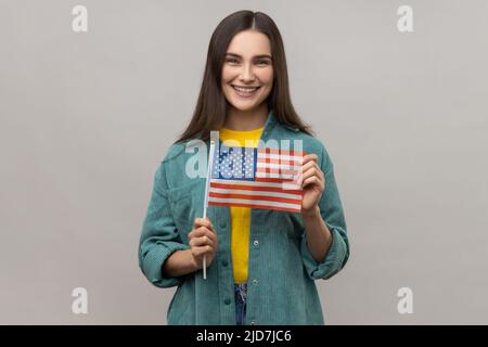 Portrait d'une jeune femme heureuse satisfaite debout et tenant le drapeau américain, célébrant les fêtes nationales, portant une veste de style décontracté. Prise de vue en studio isolée sur fond gris. Banque D'Images