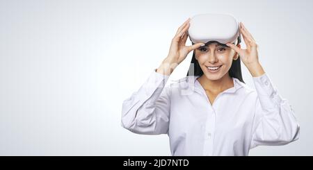 Concept de réalité virtuelle : jeune fille sourit dans une chemise légère avec des lunettes VR blanches sur sa tête sur fond gris clair avec un emplacement pour votre logo ou votre texte, Banque D'Images