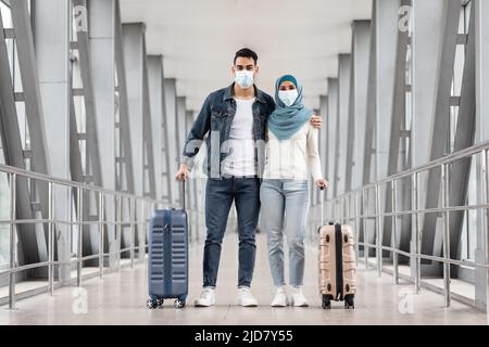 Un heureux couple musulman en masques médicaux de protection posant avec des valises à l'aéroport Banque D'Images