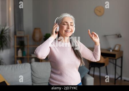Une femme européenne aux cheveux gris et aux écouteurs sans fil est heureuse d'écouter votre musique préférée et de profiter de son temps libre Banque D'Images