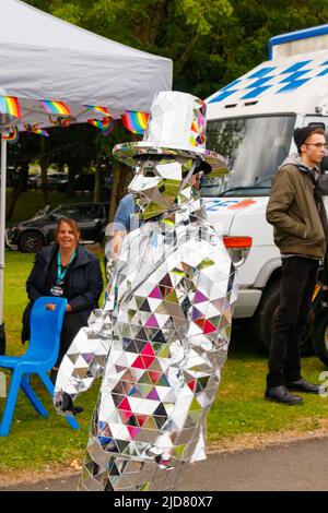 Homme et femme marchant sur pilotis avec boas en plumes arc-en-ciel et personne en costume argenté lors de l'événement Stoke gay Pride à Hanley Park le samedi 18th juin 2022 Banque D'Images