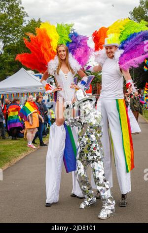 Homme et femme marchant sur pilotis avec boas en plumes arc-en-ciel et personne en costume argenté lors de l'événement Stoke gay Pride à Hanley Park le samedi 18th juin 2022 Banque D'Images