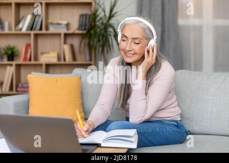 Bonne vieille femme européenne aux cheveux gris dans un casque sans fil qui travaille avec un ordinateur portable dans le salon Banque D'Images