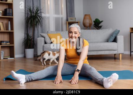 Bonne femme senior s'étirant sur un tapis de yoga, faisant des exercices de flexibilité, faisant de l'entraînement domestique dans le salon Banque D'Images