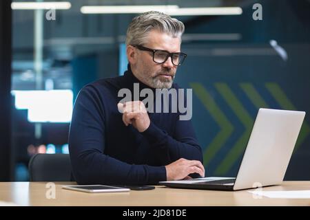 Pensant à un homme d'affaires aux cheveux gris travaillant dans un bureau moderne avec un ordinateur portable, un directeur de sexe masculin regardant l'écran d'un ordinateur portable, pensant à l'autobus de l'entreprise Banque D'Images