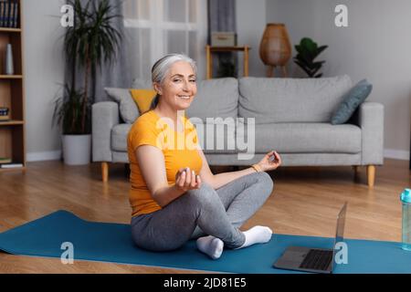 Yoga en ligne. Heureuse femme sénior méditant devant un ordinateur portable à la maison, assise en position lotus et souriante Banque D'Images