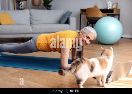 Bonne femme âgée en vêtements de sport qui s'occupe de l'exercice à la maison avec un chat et qui regarde la leçon sur un ordinateur portable Banque D'Images