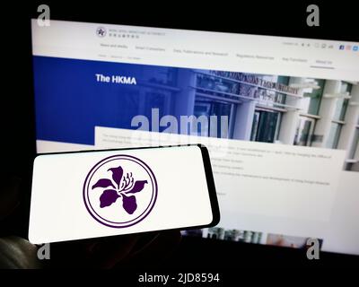 Personne tenant un téléphone portable avec le logo de l'Autorité monétaire de Hong Kong (HKMA) à l'écran en face de la page Web des affaires. Mise au point sur l'affichage du téléphone. Banque D'Images