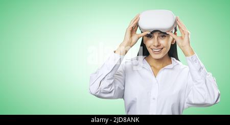 Concept de réalité virtuelle: Sourit jeune femme en chemise légère avec des lunettes VR sur sa tête sur fond vert clair avec place pour votre logo ou texte, moc Banque D'Images