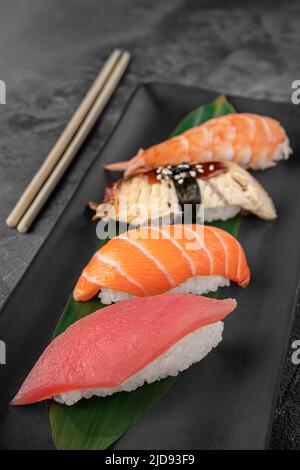 ensemble de nigiri avec crevettes tigrées, tomago, saumon, thon et feuille de bambou verte dans une assiette en céramique noire avec baguette sur fond texturé gris foncé Banque D'Images