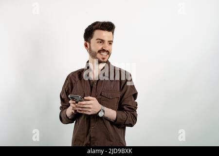 Beau jeune homme portant des vêtements décontractés posant isolé sur fond blanc aspect rêveur, en pensant tout en tenant le smartphone et en regardant de côté. Banque D'Images