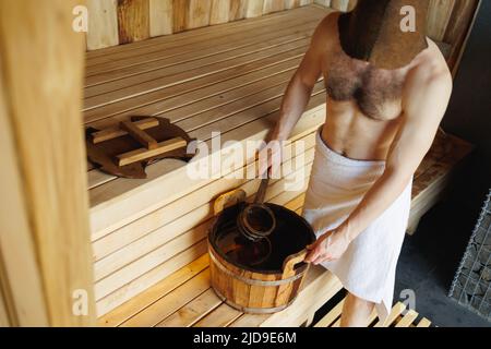 Homme qui se moque de l'eau du seau en bois dans le sauna. Homme reposant dans un bain de vapeur Banque D'Images