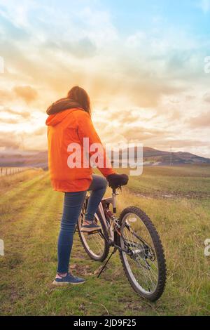 Une jeune femme hispanique portant une veste orange et un pantalon bleu se reposant sur son vélo vu de derrière au milieu du terrain rural pendant un su Banque D'Images