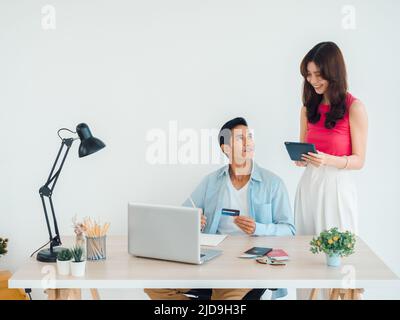 Joyeux couple asiatique, jeune homme et femme utilisant une tablette et un ordinateur portable ensemble pour la réservation de vol, la chambre d'hôtel et les informations de voyage à la maison, prêt t Banque D'Images