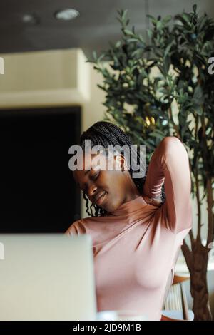 Jeune afro-américaine s'étirant le cou avec des yeux fermés inclinant de la tête vers le côté, assis et travaillant dans la maison de café. Fille noire travaillant à distance Banque D'Images