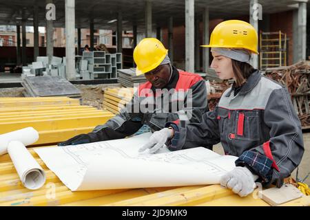 Jeune femme ingénieure confiante qui pointe sur le croquis de la construction sur un plan tout en expliquant ses détails au contremaître mature Banque D'Images