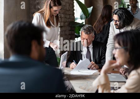 Les hommes d'affaires qui signent un contrat se sont réunis autour de la table de conférence - un homme d'affaires mature appose sa signature sur des feuilles de papier - les gens Banque D'Images