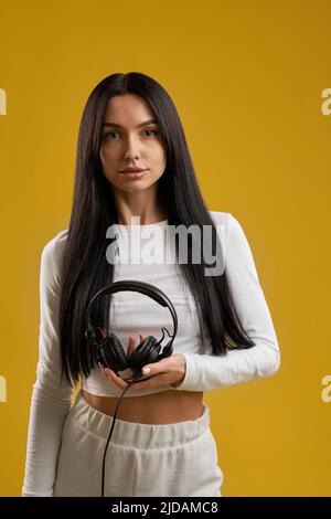 Jolie fille brune en chemise blanche tenant soigneusement le casque, tout en regardant l'appareil photo. Vue portrait d'une femme sportive avec écouteurs, isolée sur fond orange studio. Concept de musique. Banque D'Images