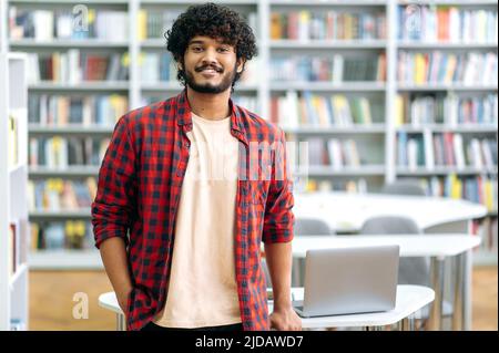 Beau homme de race mixte, indien ou arabe, en tenue décontractée, étudiant, indépendant ou enseignant, se tient dans la bibliothèque sur le fond des étagères, regarde la caméra, sourit Banque D'Images