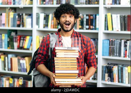 Excité heureux surpris de course mixte, étudiant, se tient dans la bibliothèque contre le fond des étagères, tient beaucoup de livres dans ses mains, regarde l'appareil photo dans la surprise, stupéfait Banque D'Images