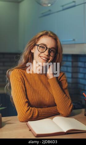 Femme aux cheveux rouges lisant un livre assis à la table dans la cuisine, souriant et regardant un appareil photo. Seul à l'intérieur. Bonne émotion. Sourire Banque D'Images