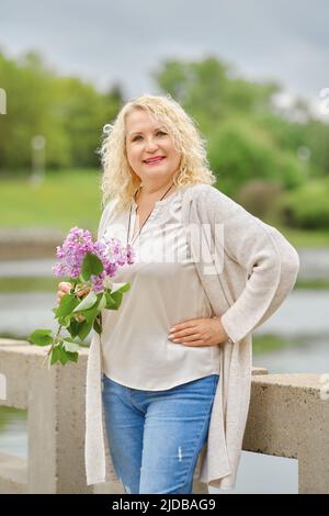 Une femme âgée gaie marche le long du remblai avec une branche de lilas dans ses mains Banque D'Images