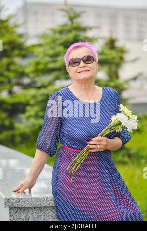 Portrait d'une femme mûre souriante aux cheveux roses courts debout près du sapin dans la rue Banque D'Images