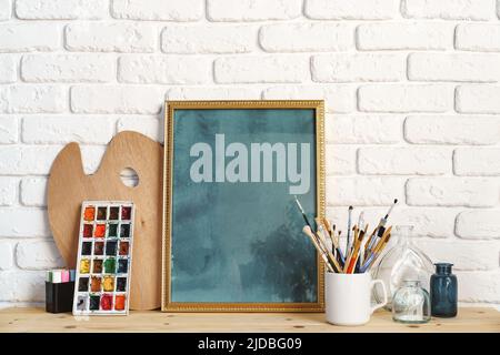 Cadre photo comme chevalet avec les outils de l'artiste sur une table en bois contre un mur en brique blanche Banque D'Images