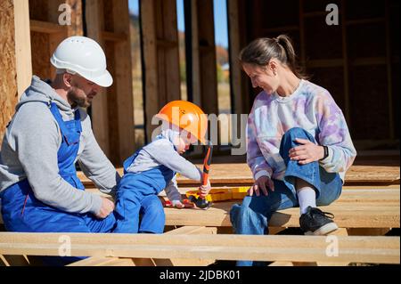 Père, mère et fils, maison en bois. Un jeune garçon aidant son papa, alors que la femme à la recherche d'eux sur le chantier de construction. Les gars portant un casque et une combinaison bleue. Menuiserie, concept de famille. Banque D'Images