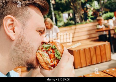 Jeune homme caucasien affamé mangeant un hamburger ou un hamburger de bœuf avec des légumes et de la sauce dans un café extérieur. Brunch le jour ensoleillé. Gros plan, fastfood, Banque D'Images