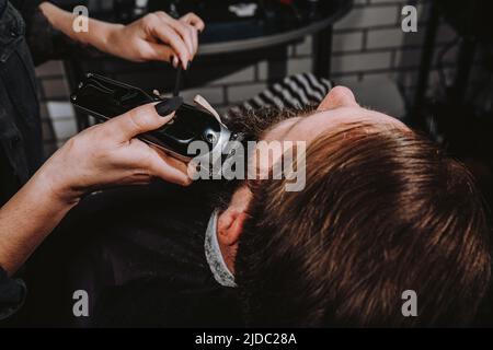 Femmes salon de coiffure professionnel servant client avec une grosse barbe épaisse par tondeuse, beau brutal jeune homme élégant obtenir une coupe de cheveux tendance, salon de coiffure noir Banque D'Images