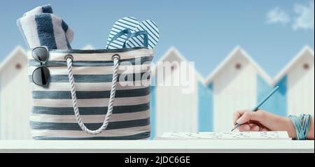 Femme se détendant à la plage et résolvant des mots croisés sur un magazine, sac de plage avec des accessoires au premier plan Banque D'Images