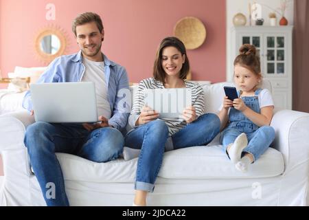 Père, mère et fille utilisant des appareils électroniques assis sur un canapé dans la salle de séjour Banque D'Images