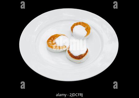 Beignets de fromage cottage Syrniki avec crème sure sur l'assiette. Arrière-plan noir isolé. Dessert caillé pour le petit déjeuner. Banque D'Images