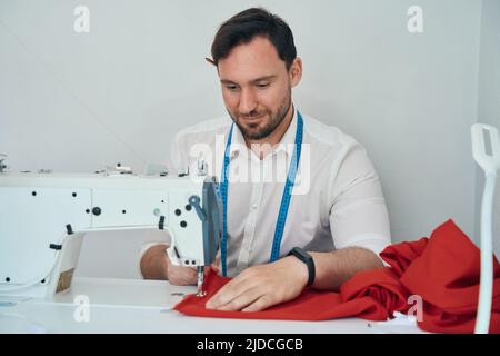 Joyeux couturier en train de créer une robe rouge avec de l'équipement Banque D'Images