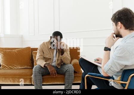 Un jeune afro-américain frustré assis sur un canapé et tenant les mains près du visage au bureau lors d'une séance avec un psychologue ou un psychiatre. Désespéré Banque D'Images