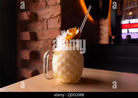 Cocktail Banana pina colada avec glace, tube à cocktail et chips d'ananas en verre avec poignée sur table en bois sur fond de brique. Bar ou café. Avant Banque D'Images