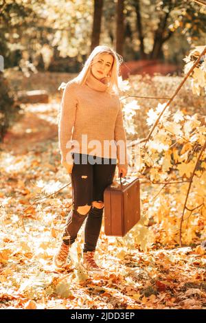 Le pull en cachemire blond en tricot féminin se tient dans le parc de la ville, tenant une vieille valise dans ses mains, le jour ensoleillé de l'automne parmi les feuilles jaunes tombées Banque D'Images